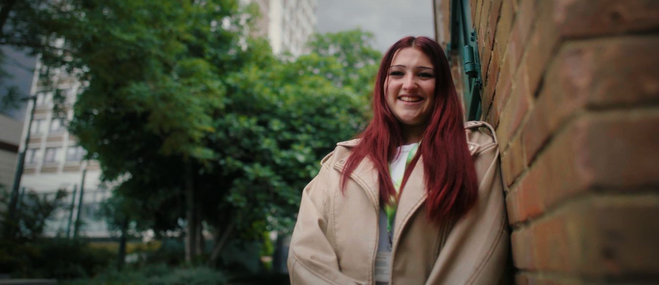 Smiling young woman with long red hair in a beige coat leans against a brick wall on a leafy city street, creating a warm, friendly mood.